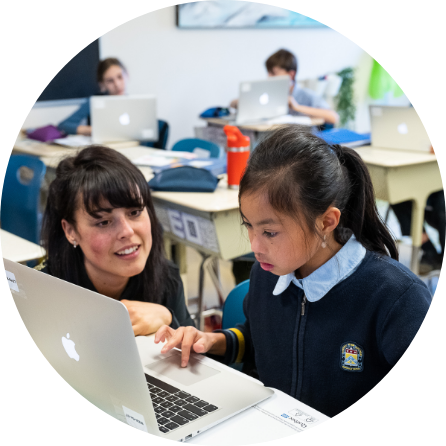 Photo d’une professeure qui aide une jeune élève dans une salle de classe. En arrière-plan, deux élèves sont assis à leur pupitre et travaillent sur leur ordinateur.
